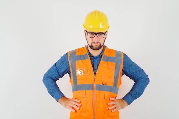 Free Photo young builder holding hands on waist in shirt, vest, helmet and looking serious. front view.