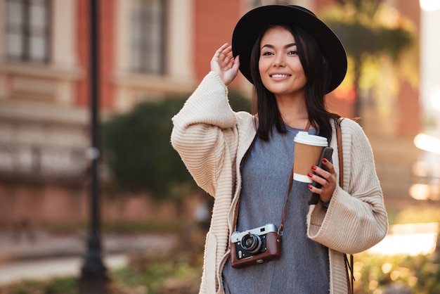 Free photo young charming asian woman in black hat holding takeaway coffee and mobile phone, looking aside while walking on street outdoor