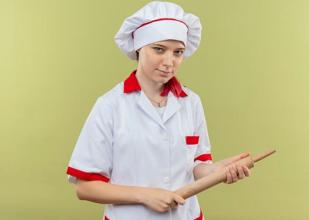 Foto gratuita il giovane chef femminile biondo fiducioso in uniforme del cuoco unico tiene il mattarello nelle mani e sembra isolato sulla parete verde