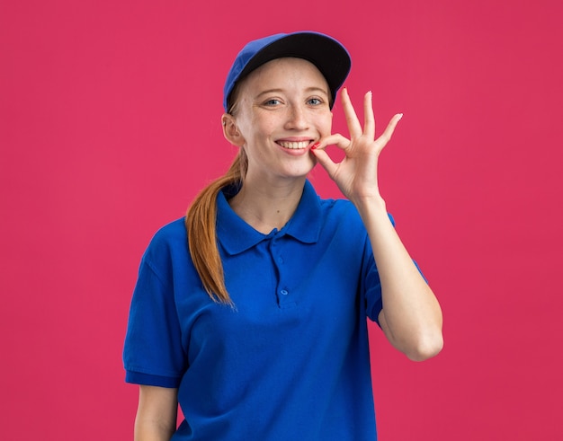 Free photo young delivery girl in blue uniform and cap   happy and cheerful making silence gesture like closing mouth with a zipper