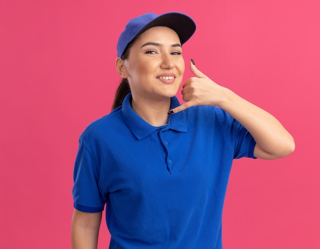 Free photo young delivery woman in blue uniform and cap looking at front smiling cheerfully making call me gesture standing over pink wall