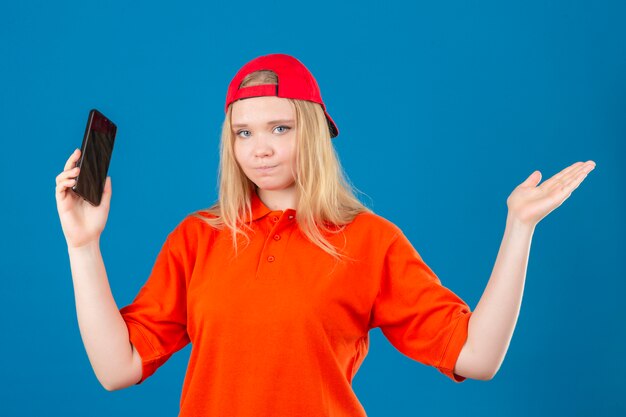 Young delivery woman wearing orange polo shirt and red cap holding smartphone shrugging shoulders and open eyes confused over isolated blue background