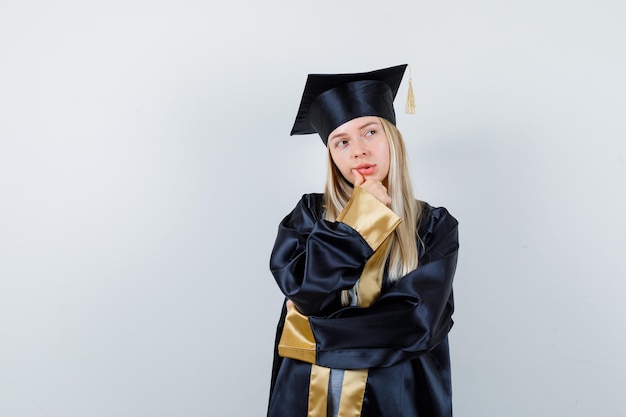 Free photo young female keeping hand on chin in graduate uniform and looking hesitant