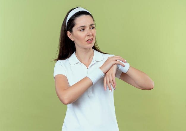 Free photo young fitness woman in headband touching her wrist looking displeased feeling pain standing over light wall