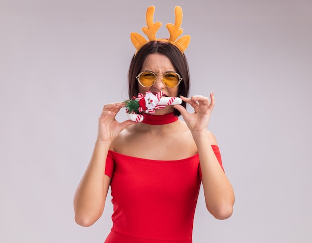 Free photo young girl wearing reindeer antlers headband and glasses holding christmas candy cane ornament near mouth biting it looking at camera isolated on white background