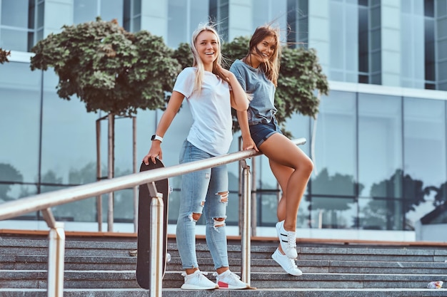 Free photo young happy hipster girls standing on steps with a skateboard on a background of the skyscraper.