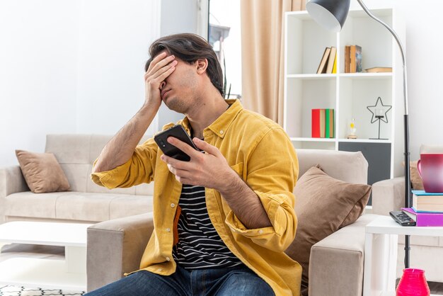 Young man in casual clothes holding smartphone looking tired and bored covering eyes with hand sitting on the chair in light living room