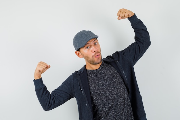 Young man showing muscles of arms in t-shirt, jacket, cap and looking confident.