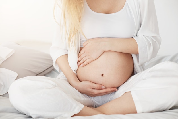 Free photo young pregnant soon to be mother touching her belly sitting in her bedroom in afternoon. pregnancy concept.