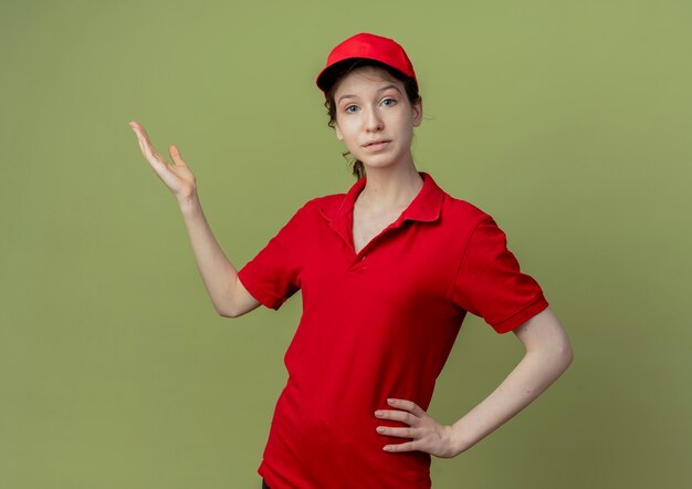 Free Photo young pretty delivery girl in red uniform and cap looking at camera putting hand on waist and showing empty hand isolated on olive green background with copy space