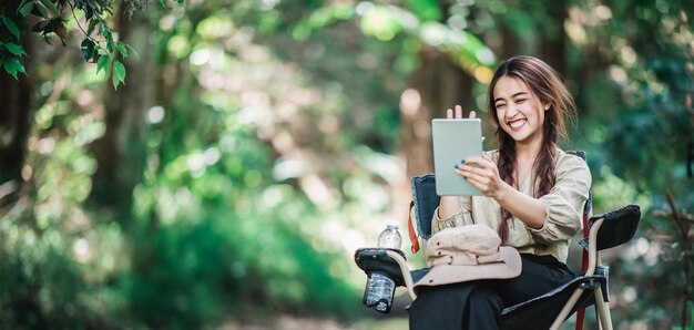 Foto gratuita giovane donna graziosa che si siede sulla sedia e usa la videochiamata del tablet mentre si accampa nello spazio della copia del parco naturale