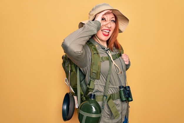 Free photo young redhead backpacker woman hiking wearing backpack and hat over yellow background doing ok gesture with hand smiling eye looking through fingers with happy face