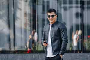 Free photo young stylish guy in glasses in black leather jacket with phone on glass background