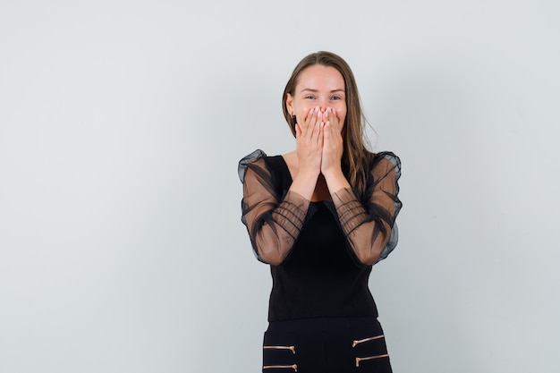 Young woman holding hands on her mouth while laughing in black blouse and looking cheerful 