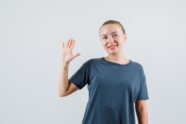 Young woman waving hand to say goodbye in grey t-shirt and looking glad