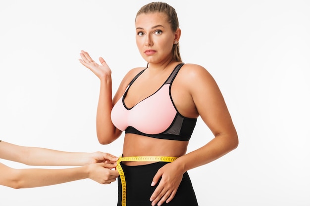 Free Photo young woman with excess weight in sporty top amazedly looking in camera while measuring waist over white background