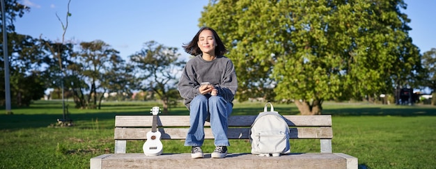 Free photo young woman with ukulele sitting on bench in park using mobile phone app holding smartphone and