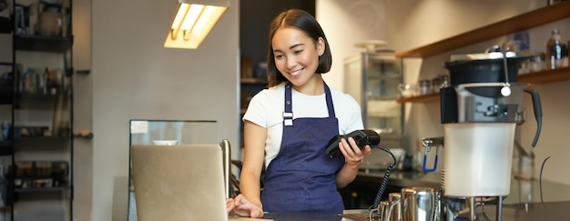 Photo gratuit bariste asiatique souriante travaillant dans un café servant un client regardant un ordinateur portable tout en utilisant un lecteur de cartes