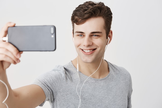 Photo gratuite beau jeune homme de race blanche aux cheveux noirs et aux yeux bleus attrayants tenant un téléphone mobile, posant pour selfie, regardant avec un sourire affectueux. beau mec faisant un appel vidéo.