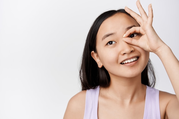 Photo gratuit beauté. jeune femme asiatique à la peau propre et éclatante, montrant ok, aucun geste sur les yeux, souriante et regardant ailleurs, debout sur blanc.