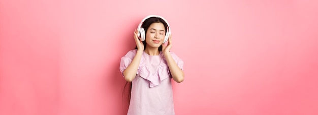 Photo gratuite belle fille asiatique ferme les yeux tout en écoutant de la musique dans des écouteurs en profitant d'un son doux debout aga