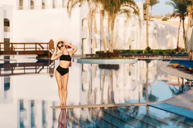 Belle jeune femme vêtue d'un élégant maillot de bain noir, bikini, fille en vacances, vacances, en station, dans un bel hôtel. Porter des lunettes de soleil élégantes, un chapeau de paille à la main.