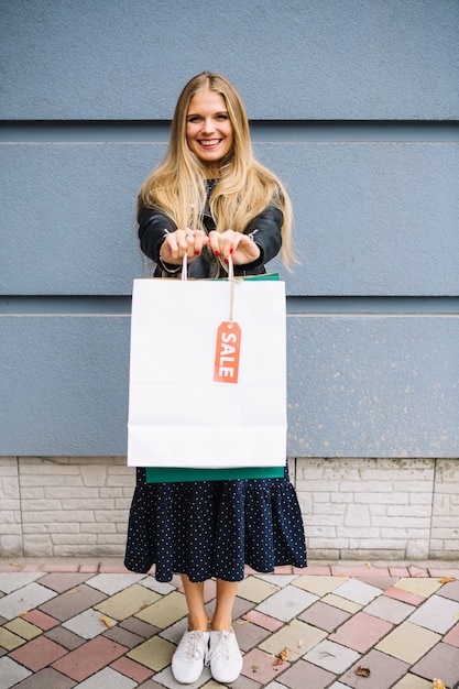 Photo gratuite blonde jeune femme debout contre le mur montrant des sacs à provisions avec étiquette de vente