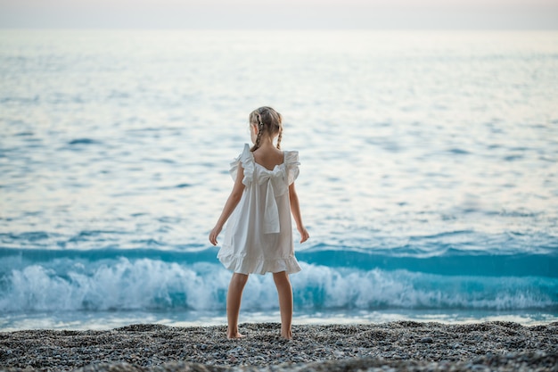 Photo gratuite bonne petite fille debout en robe blanche au bord de mer pendant le coucher du soleil.