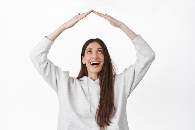 Photo gratuite concept de famille et d'assurance heureuse femme souriante regardant ses mains faisant un geste de toit de maison sur le toit debout optimiste sur fond blanc