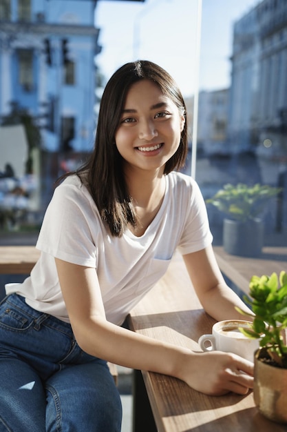 Photo gratuit concept de loisirs et de style de vie urbain pour les jeunes sourire jolie et jolie fille asiatique avec une courte coupe de cheveux sombre s'asseoir avec désinvolture café près des fenêtres derrière les rues de la ville boire du café en discutant