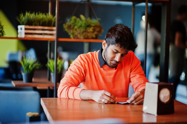 Photo gratuite confiant jeune homme indien en tenue décontractée intelligente comme un pull orange assis au café et utilisant son téléphone portable