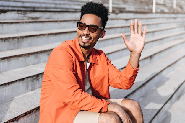 Photo gratuite cool homme brun heureux à la peau foncée dans des lunettes de soleil et une veste orange sourit, salue la main et s'assoit dans les escaliers