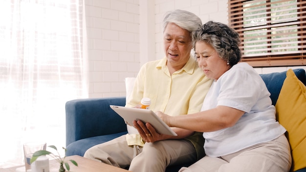 Photo gratuite couple de personnes âgées asiatique à l'aide de la tablette recherche des informations sur les médicaments dans le salon, couple utilisant le temps ensemble en position couchée sur le canapé lorsque détendu à la maison.