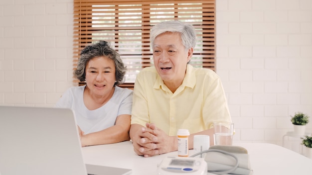 Photo gratuite couple de personnes âgées asiatique utilisant la conférence de l'ordinateur portable avec médecin sur les informations de médecine dans le salon, couple utilisant le temps ensemble en position couchée sur le canapé à la maison.