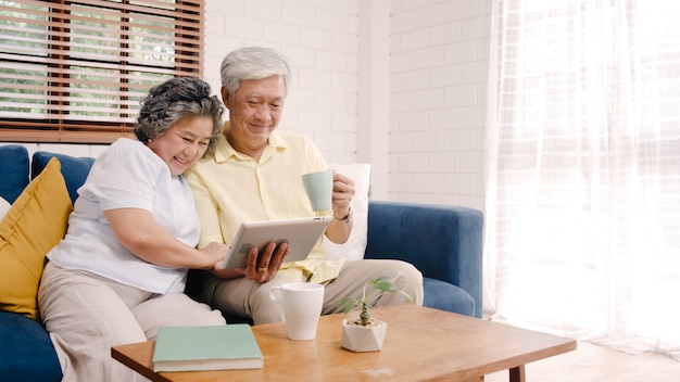 Photo gratuite couple de personnes âgées asiatiques utilisant une tablette et buvant du café dans le salon à la maison, un couple profitant d’un moment d’amour en position couchée sur un canapé pour une détente à la maison.