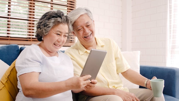 Photo gratuite couple de personnes âgées asiatiques utilisant une tablette et buvant du café dans le salon à la maison, un couple profitant d’un moment d’amour en position couchée sur un canapé pour une détente à la maison.