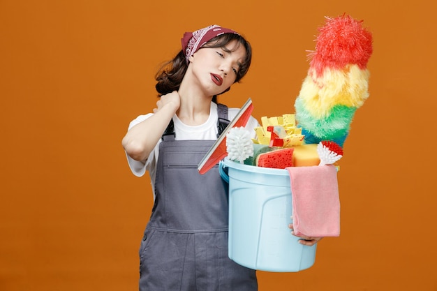 Photo gratuite fatigué jeune femme nettoyeur en uniforme et bandana tenant un seau d'outils de nettoyage en gardant la main sur le cou en regardant le côté isolé sur fond orange