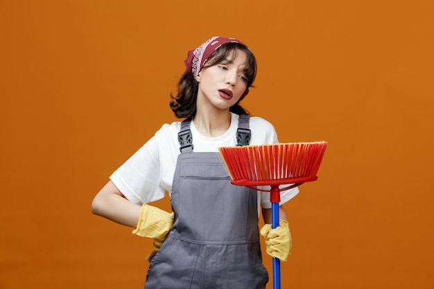 Photo gratuite fatigué jeune femme nettoyeuse portant des gants en caoutchouc uniformes et un bandana tenant une vadrouille à raclette en le regardant tout en gardant la main sur la taille isolée sur fond orange