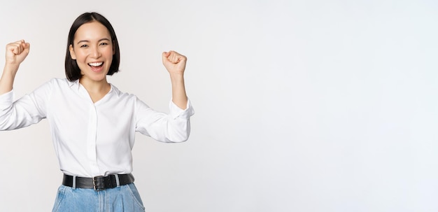 Photo gratuit femme asiatique enthousiaste se réjouissant de dire oui à l'air heureux et célébrant la victoire championne danse poing pompe geste debout sur fond blanc