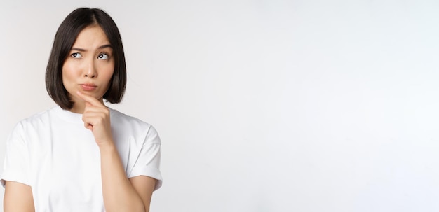 Photo gratuite une femme asiatique réfléchie qui regarde de côté en pensant à faire des suppositions en pensant ou en choisissant qch debout sur fond blanc