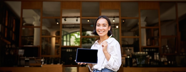 Photo gratuite une femme asiatique souriante montrant un écran de tablette numérique propriétaire d'un café montrant smth debout devant le café