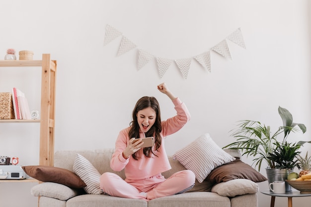 Photo gratuite femme bouclée en costume rose se réjouit du jeu gagnant sur smartphone