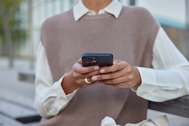 Photo gratuite femme dans des vêtements élégants tient un téléphone mobile surfe sur internet sans fil fait des achats en ligne est assise sur un banc à l'extérieur connecté à internet sans fil communique à distance