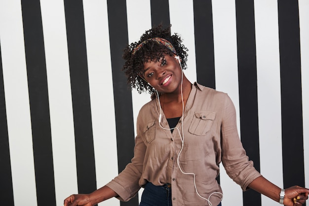 Photo gratuite femme écoutant de la musique dans les écouteurs et dansant dessus. fille afro-américaine souriante se tient dans le studio avec des lignes verticales blanches et noires à l'arrière-plan