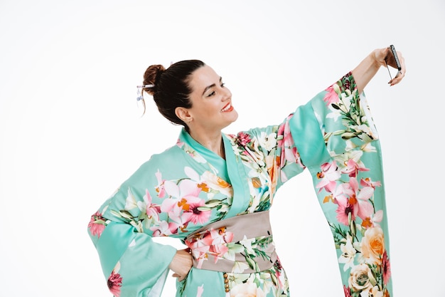 Photo gratuite femme en kimono japonais traditionnel souriant heureux et positif faisant gaiement selfie à l'aide de smartphone sur blanc