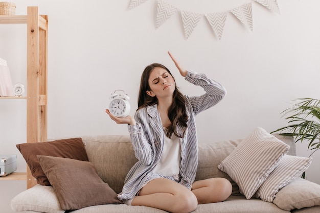 Photo gratuit la femme a mal à la tête. femme en pyjama posant dans le salon avec réveil