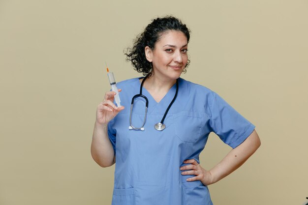 Femme médecin d'âge moyen heureuse et confiante portant un uniforme et un stéthoscope autour du cou tenant une seringue avec une aiguille regardant la caméra tout en gardant la main sur la taille isolée sur fond olive