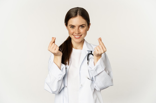 Photo gratuit femme médecin montrant des coeurs de doigt souriant avec soin debout en uniforme sur fond blanc