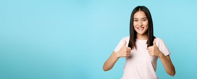 Photo gratuite fille asiatique enthousiaste montrant les pouces vers le haut comme et approuvant souriante satisfaite debout sur le dos bleu