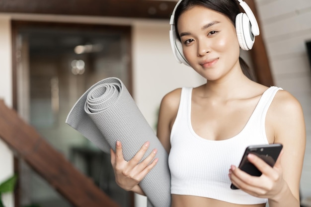 Photo gratuit une fille asiatique souriante dans un casque, écoutant de la musique à la maison et faisant de l'exercice avec un tapis de sol tenant un p...